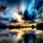 Nature landscape profile photo: dramatic sunset over a calm lake with mirrored storm clouds and a silhouetted treeline in vivid blues and golds.