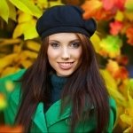 Profile photo of a beautiful woman wearing a black beret and green coat, with vibrant autumn leaves in the background.