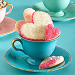 A heart-shaped cookie with white and pink icing resting on a blue teacup, accompanied by another cookie and pastel teacups in the background, used as a forum avatar.