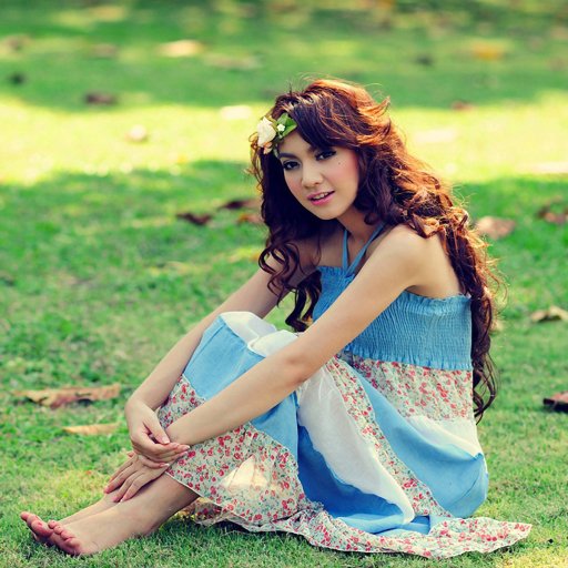 Profile avatar of a brunette woman with long hair in a blue sundress, barefoot with her feet visible, sitting on grass and smiling while hugging her knees.