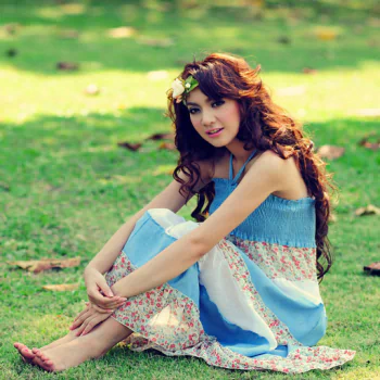 Profile avatar of a brunette woman with long hair in a blue sundress, barefoot with her feet visible, sitting on grass and smiling while hugging her knees.