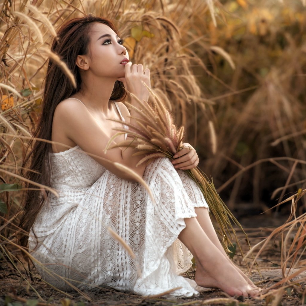 Forum avatar of an Asian woman and model with long brunette hair in a white lace dress, seated barefoot among tall grasses, gazing thoughtfully while holding dried reeds.