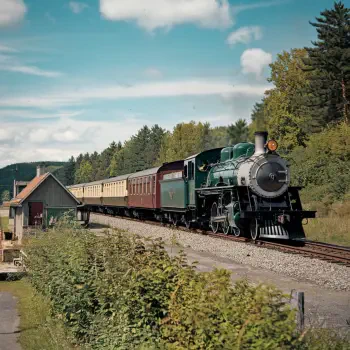 A vintage green locomotive passes a quaint station, pulling a set of wooden passenger cars along a scenic railway surrounded by lush trees and a clear sky.