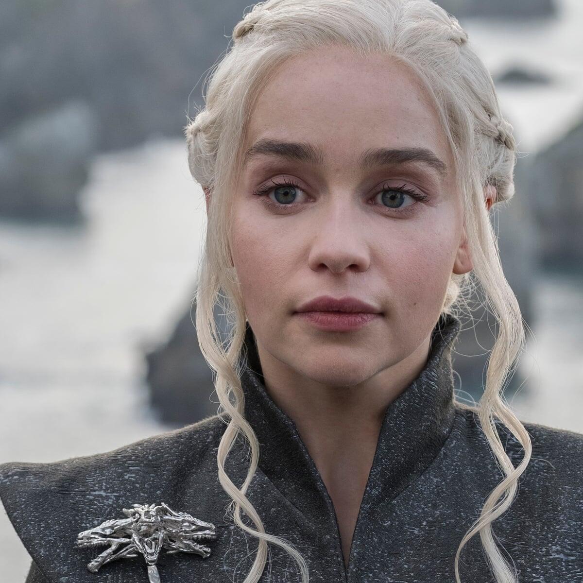 A close-up of Daenerys Targaryen, portrayed by Emilia Clarke, from Game of Thrones. She has platinum blonde hair, wearing a dark outfit, with a serious expression against a scenic backdrop.