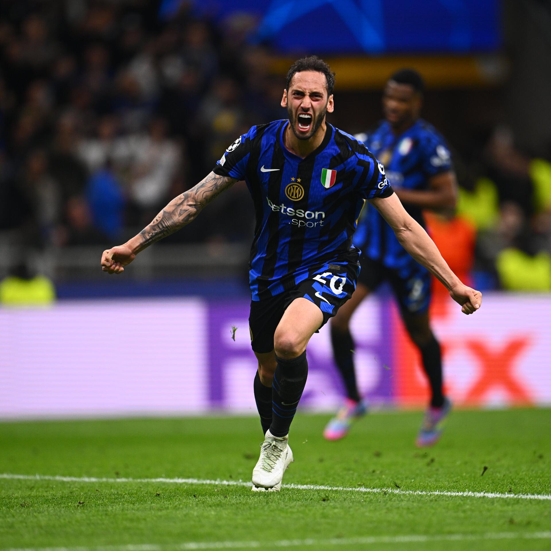 Hakan Çalhanoğlu celebrating a goal wearing Inter Milan's blue and black soccer jersey on the field.