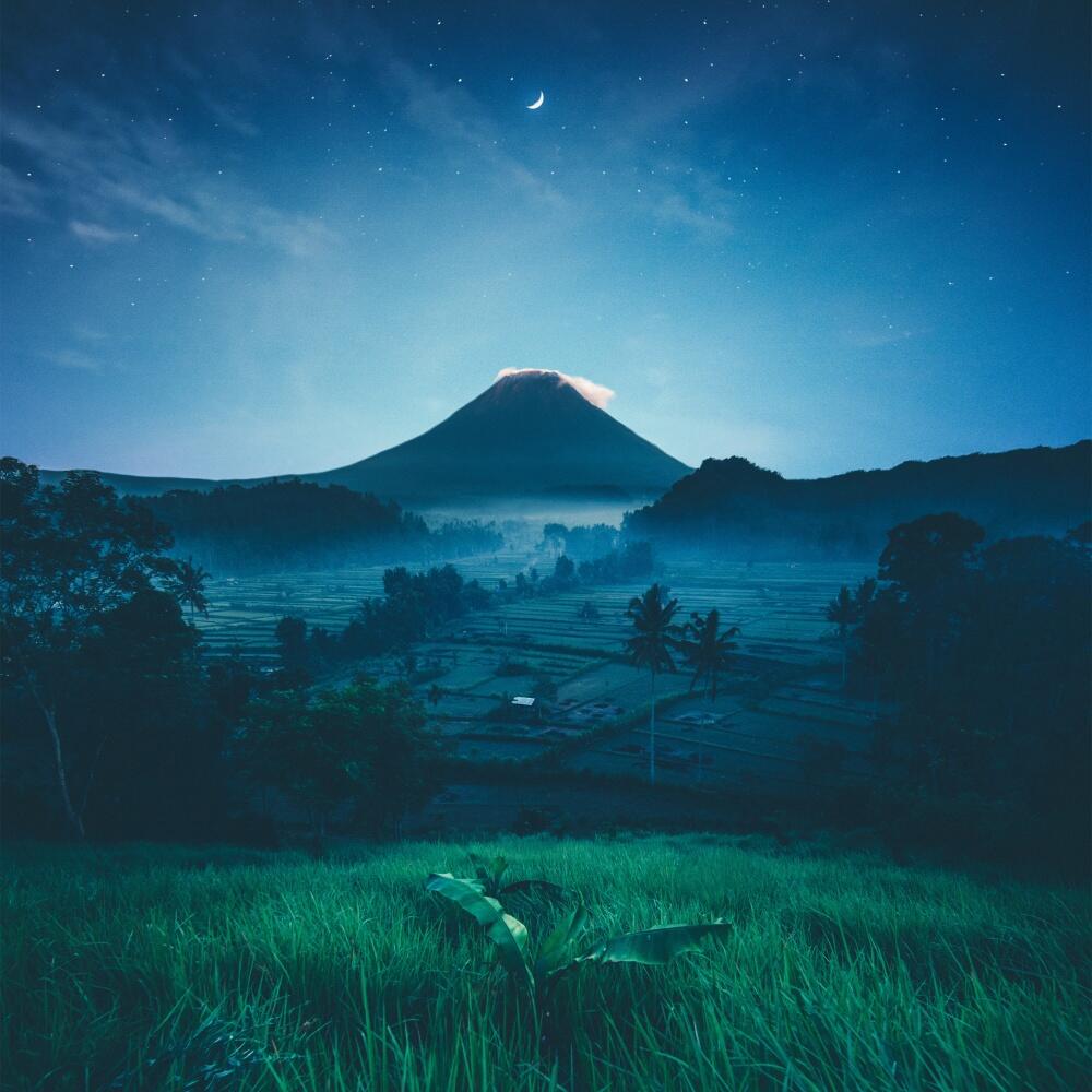 Forum avatar: night nature scene with grassy foreground, misty valley and distant silhouetted volcano beneath a crescent moon and starry sky.