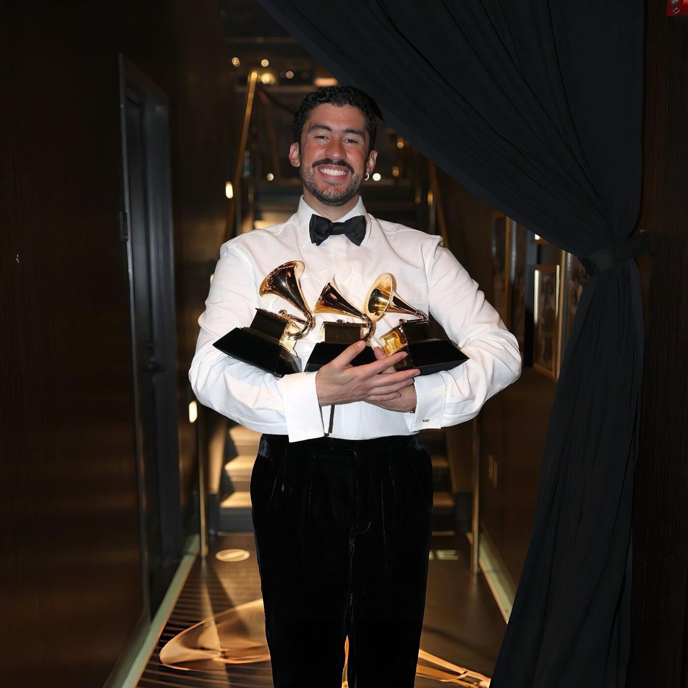 Forum avatar of a smiling Latin music artist in a tuxedo holding three Grammy Awards on a dimly lit corridor.