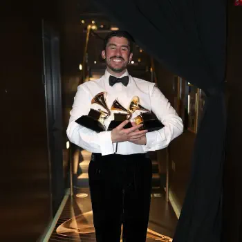 Forum avatar of a smiling Latin music artist in a tuxedo holding three Grammy Awards on a dimly lit corridor.