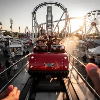 Forum avatar/profile photo of a red roller coaster car hurtling toward the viewer, riders cheering as carnival lights and a Ferris wheel glow at sunset.