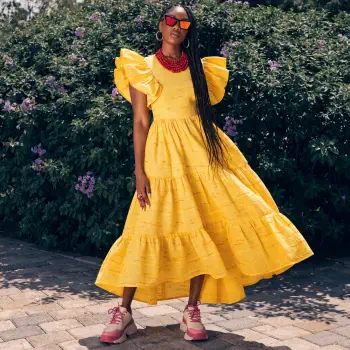Forum avatar/pfp of a woman in a bright yellow tiered dress with oversized ruffled sleeves and a red necklace, wearing sunglasses and long braids, standing outdoors in chunky pink sneakers.
