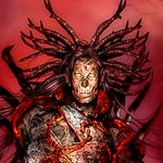 Dark warrior with a fierce, intricate mask and wild, spiked headdress, set against a deep red background, embodying a powerful and intense presence.