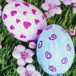 Decorated Easter eggs in pink and blue patterns sit on green grass surrounded by spring flowers, capturing the cheerful essence of springtime.