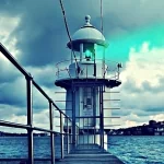 A striking lighthouse stands at the end of a wooden pier, surrounded by water and dramatic clouds, making this an engaging profile photo.